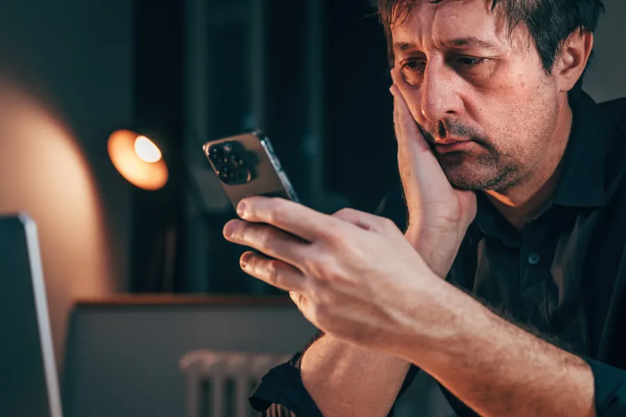 Homem de expressão preocupada olhando para o celular à noite, com o rosto apoiado na mão.